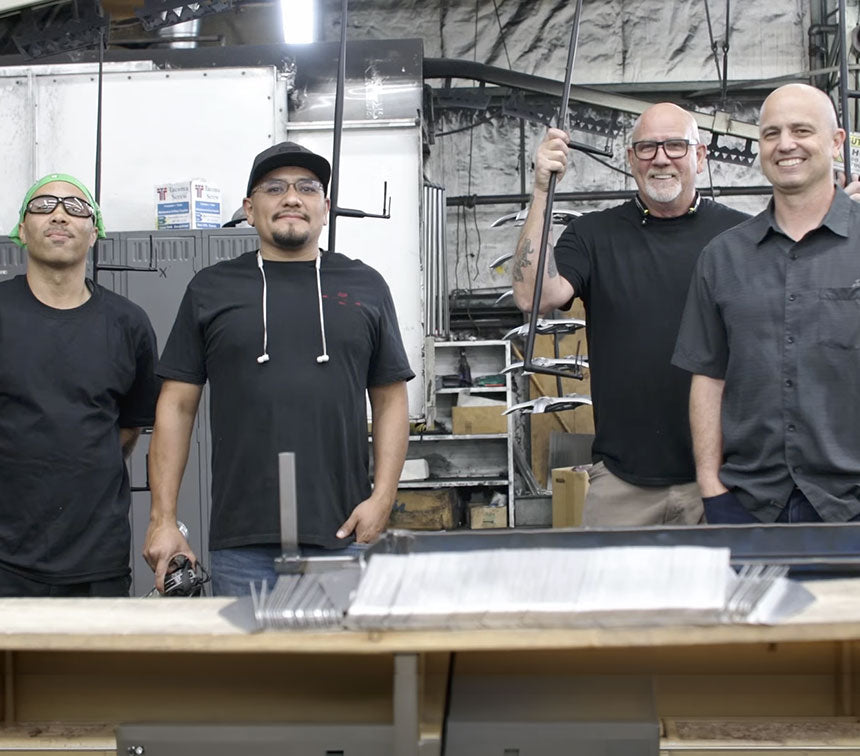 Four men standing together in a workshop or industrial interior, with metal racks, tools, and a workbench in the background. Two men are wearing black shirts, one wears a dark gray button-up shirt, and one wears a black t-shirt with a cap and glasses.