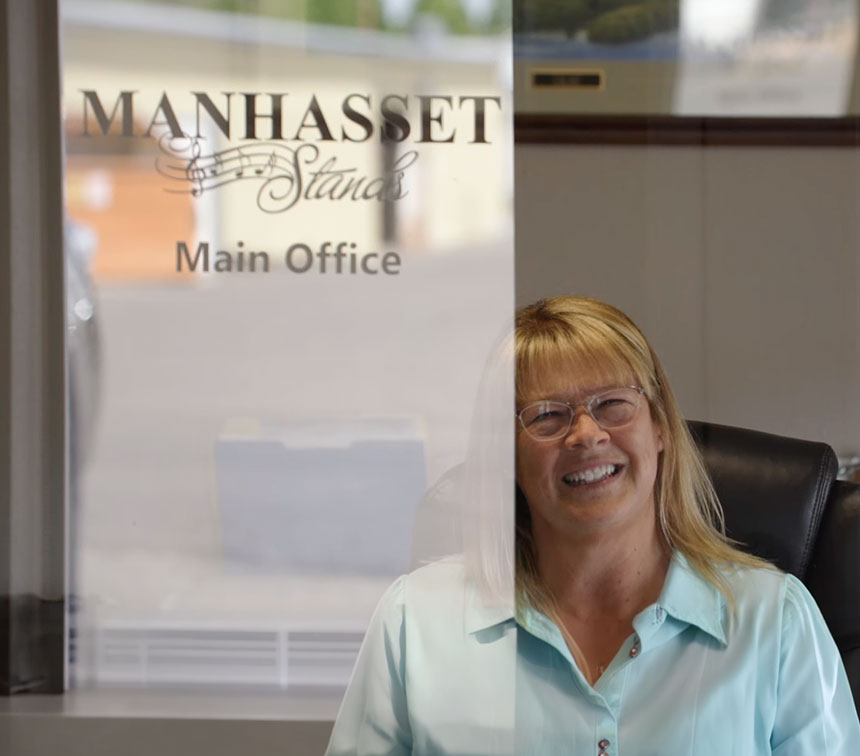 A smiling blonde woman wearing glasses and a light blue collared shirt sits in a black office chair in front of a glass door with 'MANHASSET Stands Main Office' printed on it.