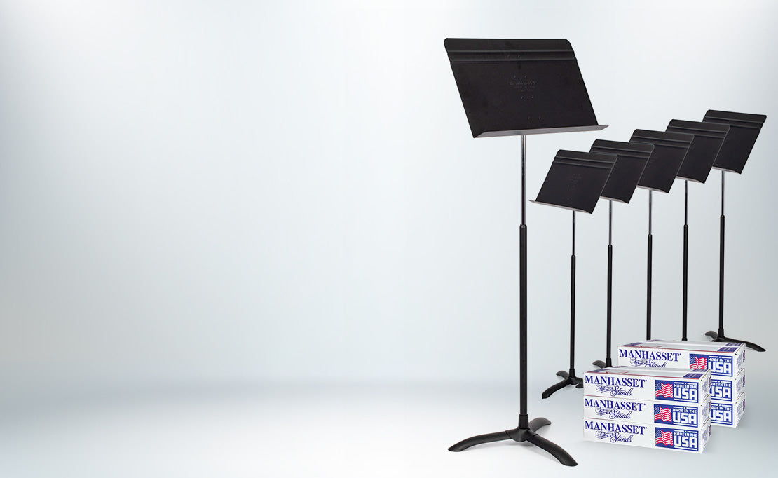 Five black music stands arranged in a row on a light gray studio background, with several white and blue Manhattan Sand & Steel boxes stacked underneath the front stands.