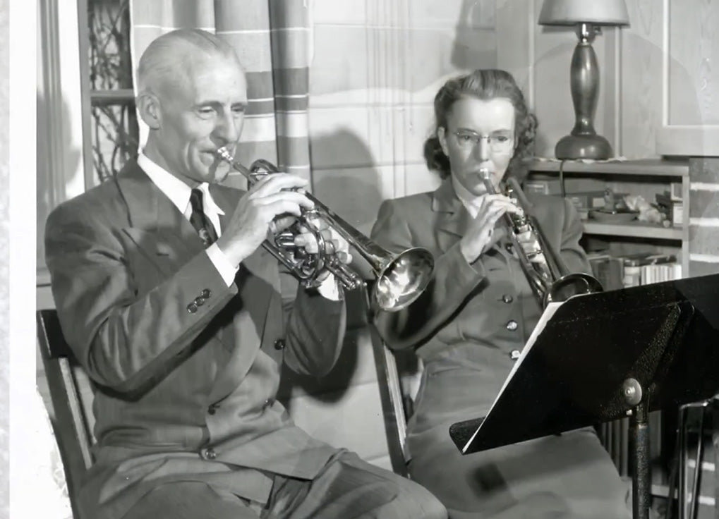 Old black and white photo from the early-mid 20th century showing two seated musicians playing a cornet and trumpet in front of a Manhasset stand.