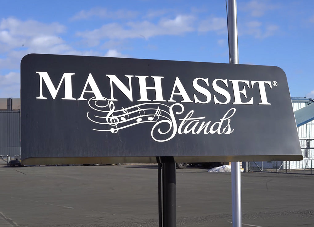 A black rectangular sign with white lettering that reads "MANHASSET Stands" and a music note graphic, mounted on a pole outdoors under a blue sky.