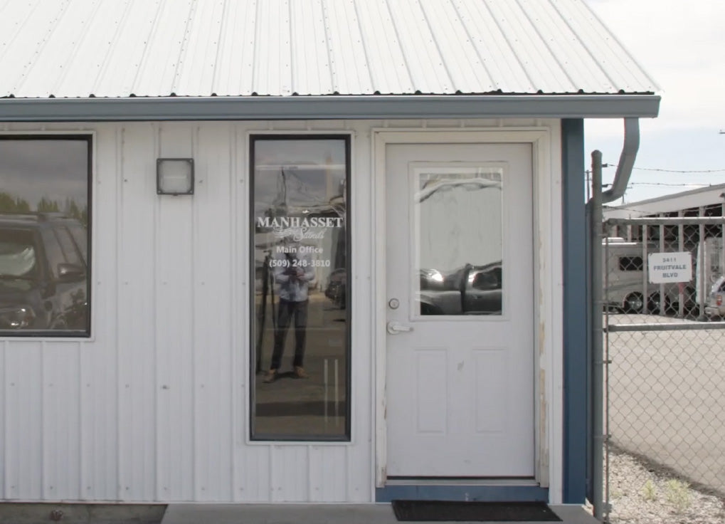 A small white building with a metal roof, featuring a white door with a window, a tall window on the left and a Manhasset logo. A chain-link fence is visible on the right side.