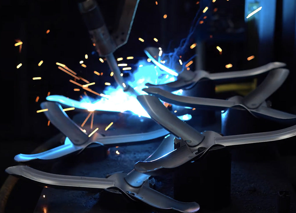 A close-up action shot of a welding torch generating a bright blue-white arc and orange sparks while holding several curved silver metal pieces in a clamp setup.