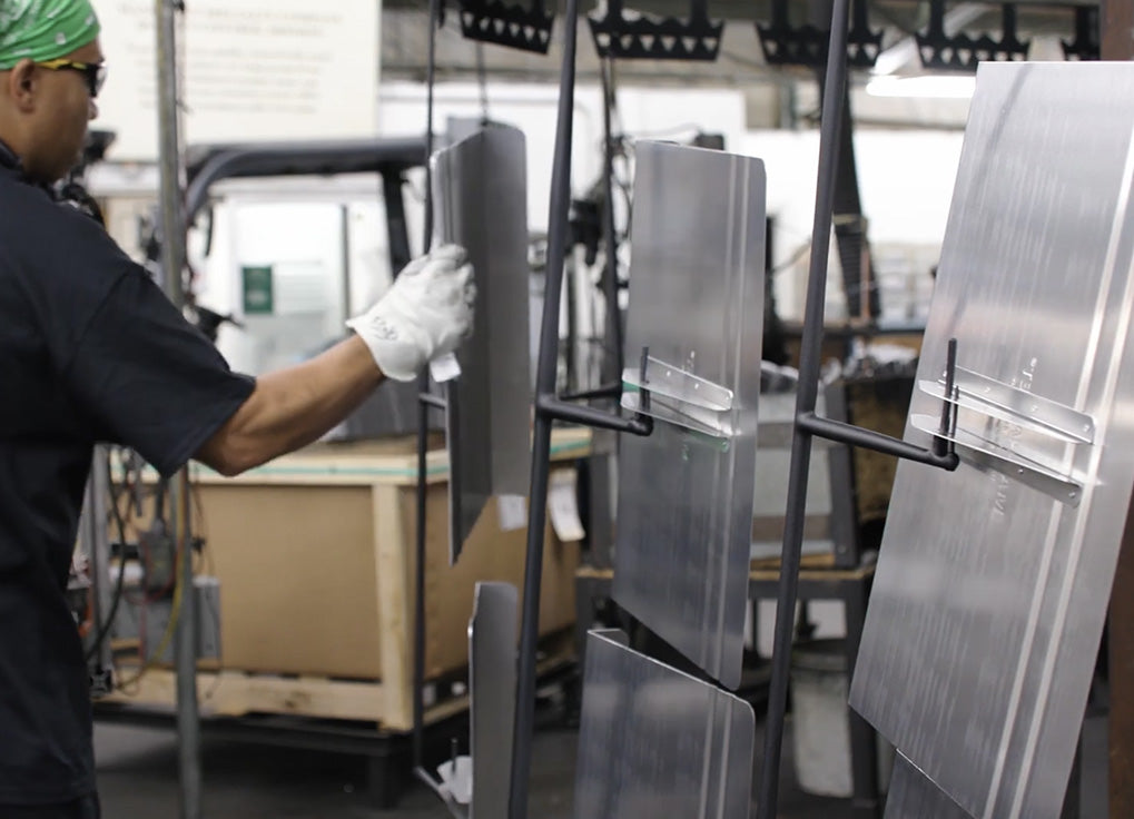 A worker wearing a green headwrap, black shirt, and white gloves handles a large brushed metal panel in a manufacturing or fabrication facility.