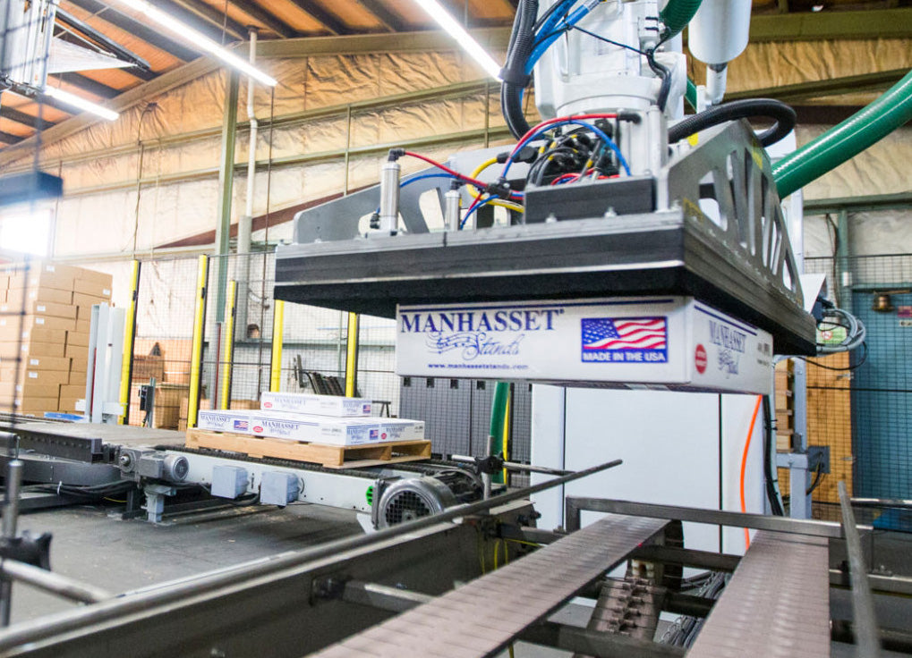 A large MANHASSET industrial robotic arm and pallet transfer system in a warehouse setting, with a pallet loaded with boxes positioned under the robotic gripper.