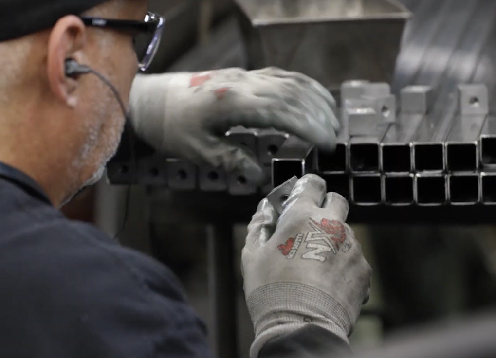 A close-up shot of a worker wearing protective gloves and glasses, handling a row of square metal pipes in an industrial setting.