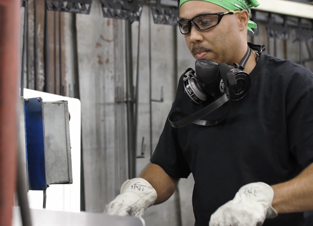 A man wearing a black t-shirt, white work gloves, and a green bandana stands in an industrial workshop environment, looking down at a task with a respirator around his neck.