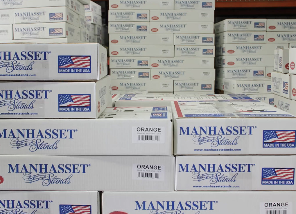 Stacks of white MANHASSET Stands orange soda beverage boxes on a store shelf, with visible MANHASSET branding, 'Made in the USA' flags, and 'Orange' flavor labels.