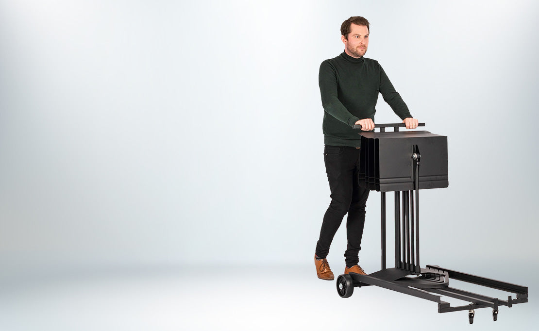 A man in a dark green turtleneck sweater and black pants stands beside a black wheeled Harmony stand trolley with a top-mounted monitor, photographed against a clean light grey studio background.