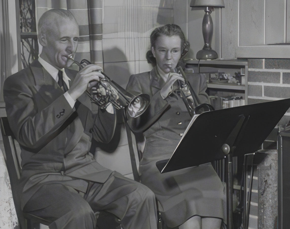 Black and white photo of a man and a woman seated indoors, each holding a trumpet near a music stand.