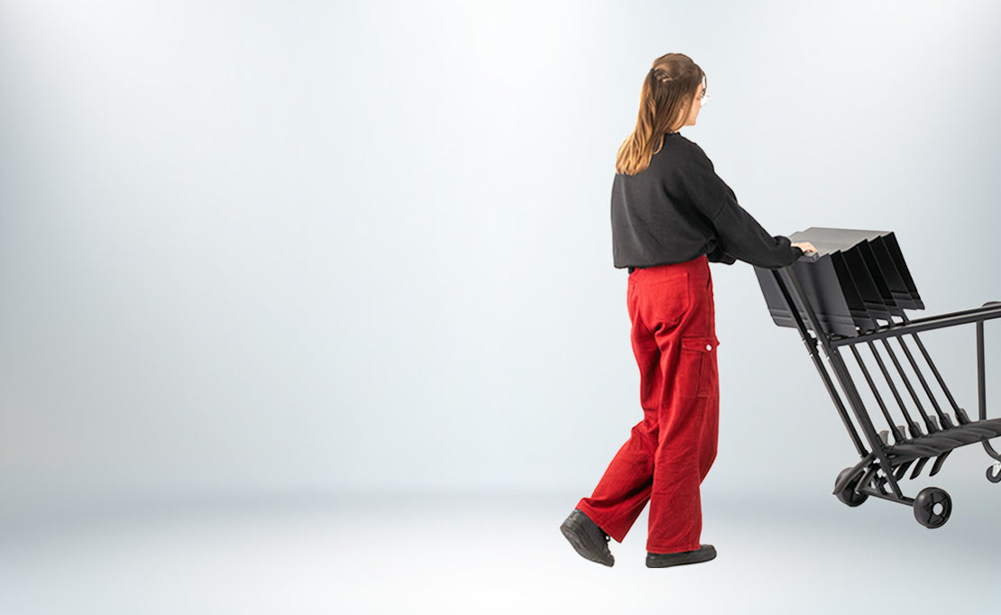 A woman seen from behind wearing a black long-sleeve top and red pants, pushing a black metal Manhasset stand cart against a light gray studio background.