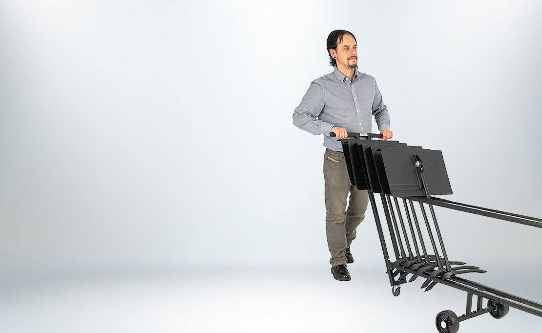 A man in a gray shirt and brown pants pushes a black metal Symphony stand cart for up to 25 stands against a light grey studio background.