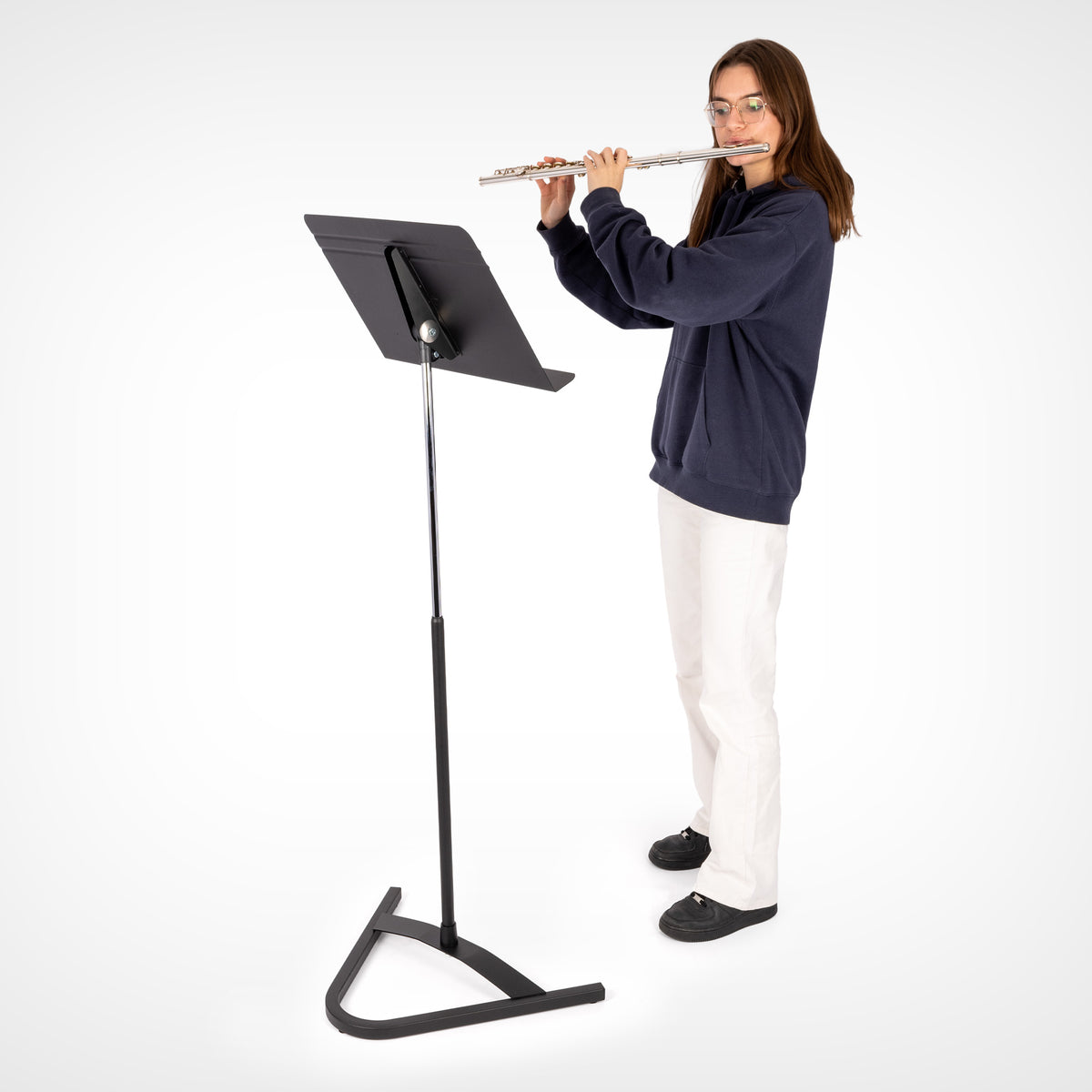A woman playing a silver flute in front of a black Harmony stand on a white studio background.