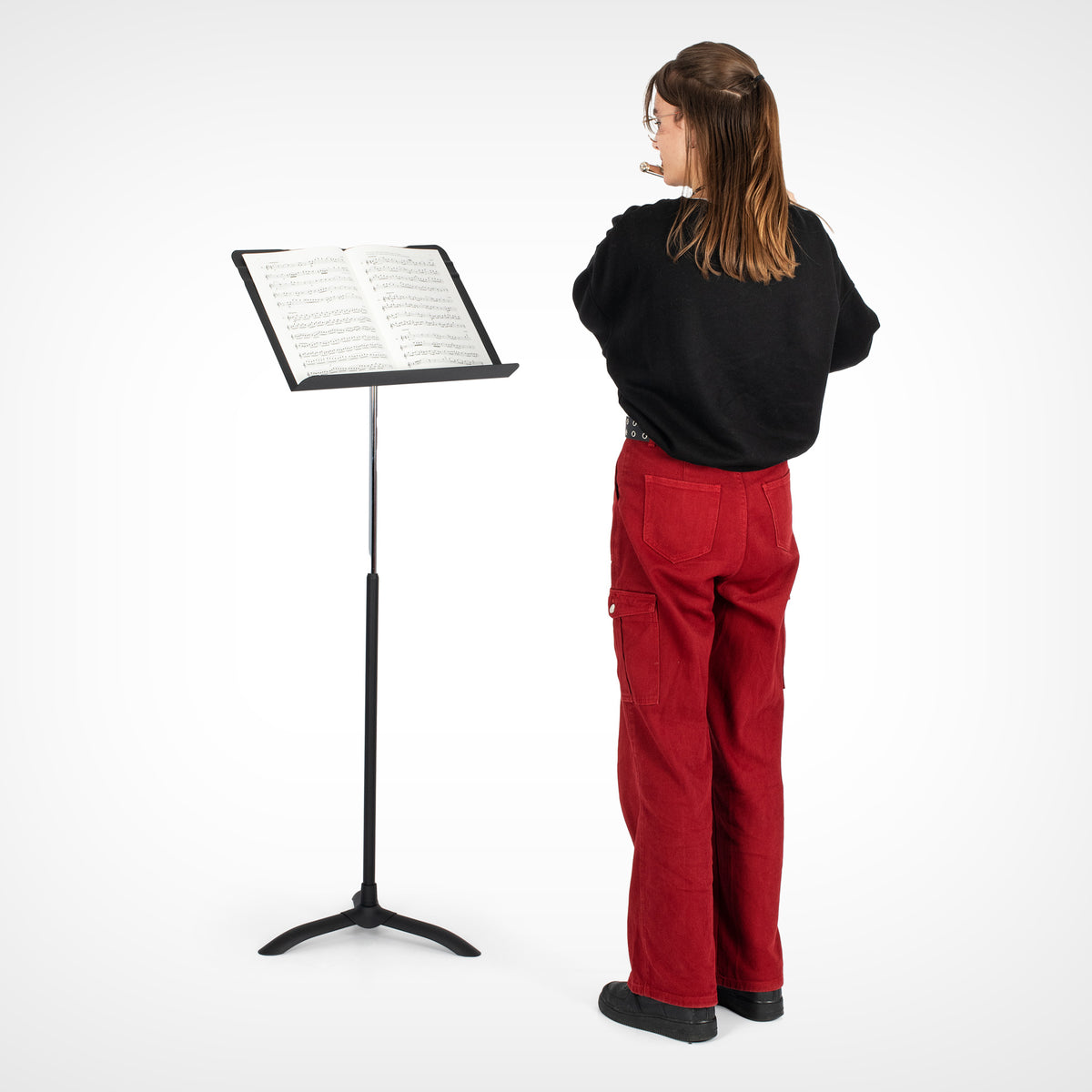 A person stands with their back to the camera next to a black adjustable music stand holding sheet music, wearing a black long-sleeve top and red cargo pants against a white studio background.